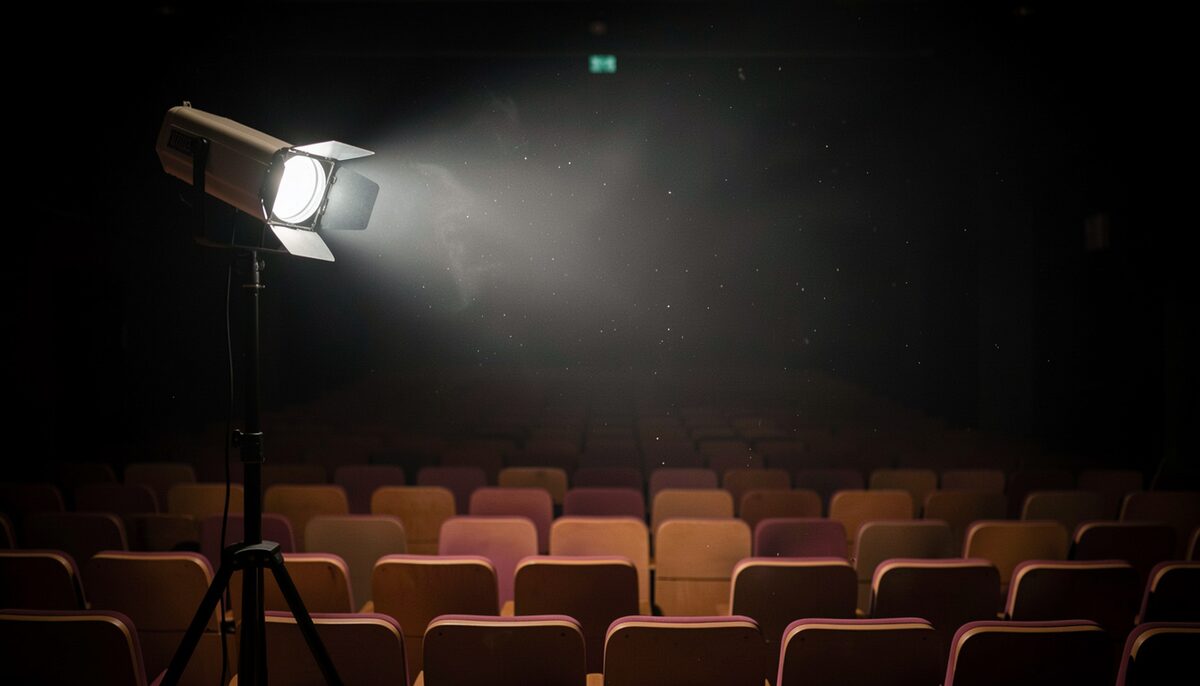 A theater stage at night lit by a single work light on a stand, empty seats stretching into darkness, dust in the beam, no text