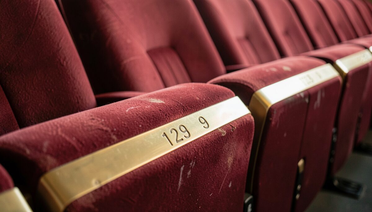 A row of theater seat numbers in brass on armrests, close up, velvet seats slightly worn, warm house light, no people